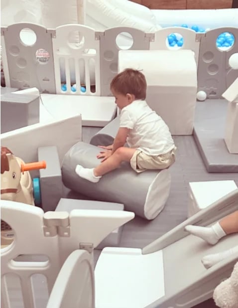 Toddler in white shirt sitting on gray toy car inside an indoor play area with white and gray play structures