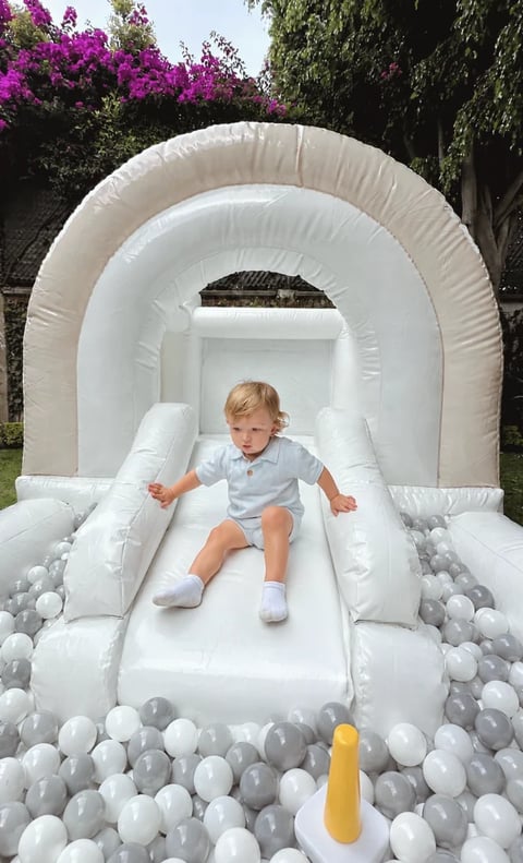 Toddler sliding down a white inflatable bounce house slide filled with gray and white plastic balls in a garden setting