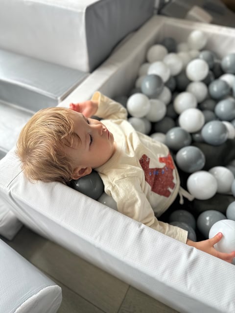 Toddler lying in a ball pit filled with gray and white balls, wearing a cream-colored jacket