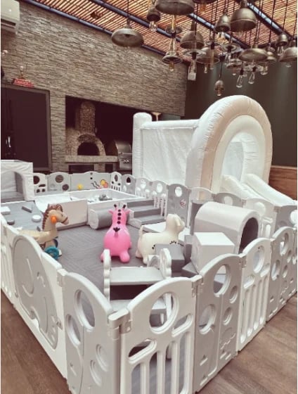 Modern playpen setup with white plastic gates, toys, and furniture in an industrial loft space with brick walls and pendant lights