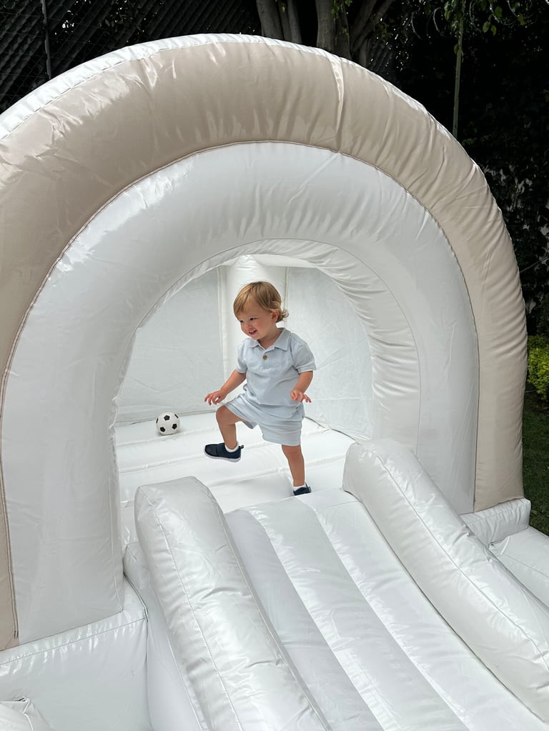 Toddler playing on a white inflatable bounce house slide with a soccer ball indoors