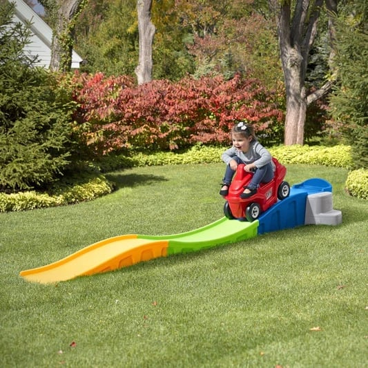 Toddler in red toy car on grass with yellow and green ramp in backyard with trees and flowering shrubs