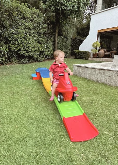 Toddler in red shirt riding a red and green toy car on a colorful ramp in a grass backyard
