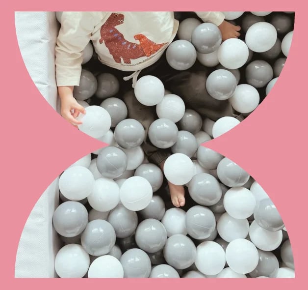 Child sitting in hourglass-shaped ball pit with white and gray plastic balls on pink background