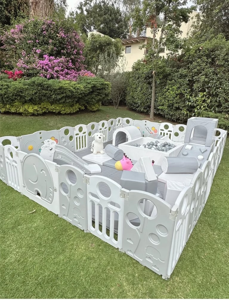 White modular baby playpen with interactive panels set up on grass in a garden surrounded by colorful flowers and trees